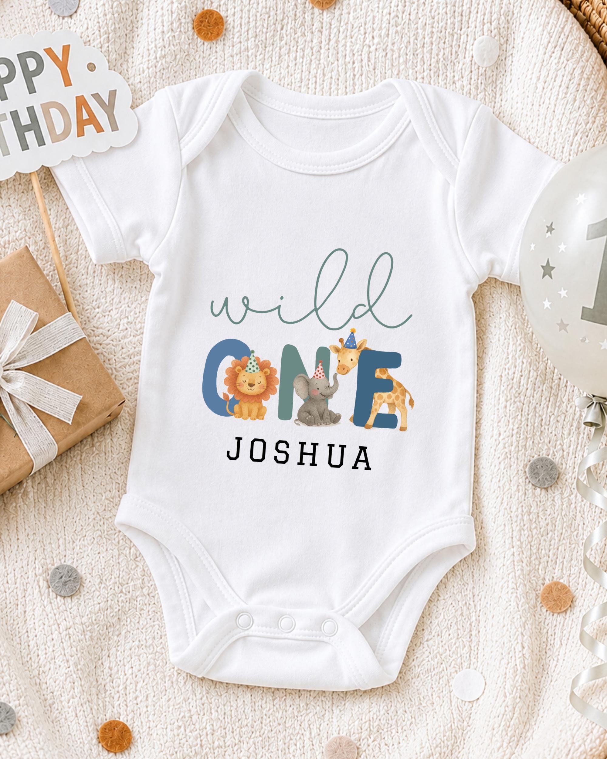 A white onesie with the text "wild one Joshua" printed on it, along with illustrations of a lion, giraffe, and elephant. The onesie is laid out on a blanket, surrounded by a gift box, a birthday balloon, and confetti.