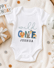 A white onesie with the text "wild one Joshua" printed on it, along with illustrations of a lion, giraffe, and elephant. The onesie is laid out on a blanket, surrounded by a gift box, a birthday balloon, and confetti.