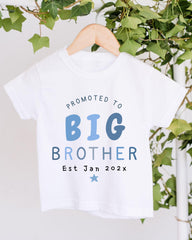 A white t-shirt with text that reads "PROMOTED TO BIG BROTHER Est Jan 202x" hanging on a wooden hanger.