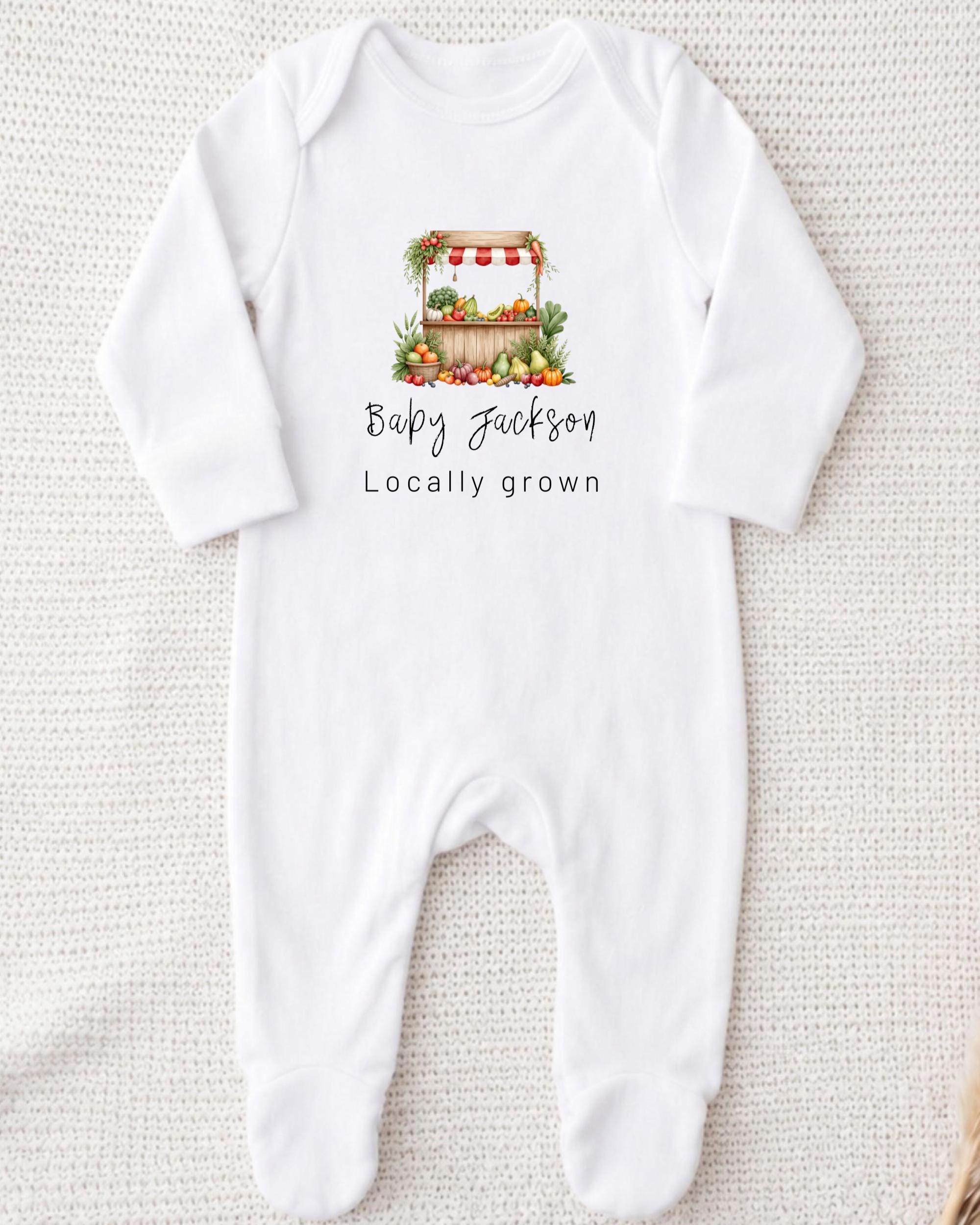 A white onesie with a printed design of a fruit stand and the text "Baby Jackson Locally grown" printed on it.