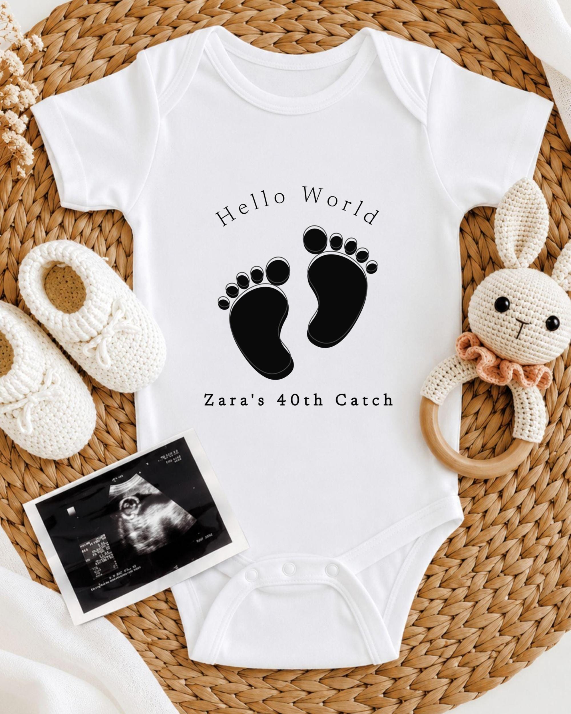 A baby onesie with the text "Hello World" and "Zara's 40th Catch" printed on it, along with a pair of white crocheted shoes and a stuffed rabbit.