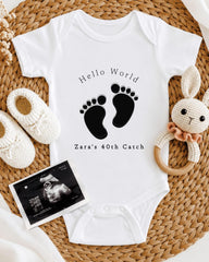 A baby onesie with the text "Hello World" and "Zara's 40th Catch" printed on it, along with a pair of white crocheted shoes and a stuffed rabbit.