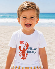 A young boy with a big smile on his face, wearing a white t-shirt with a lobster graphic and the text "Feelin' Snappy" printed on it, standing on a sandy beach with the ocean in the background.