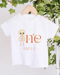 A white t-shirt with a skeleton design and the text "One - Emily" printed on it, hanging on a wooden hanger.