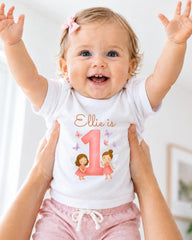 A smiling baby girl wearing a white t-shirt with a personalized number 1 and fairy characters, being held up by someone's arms.