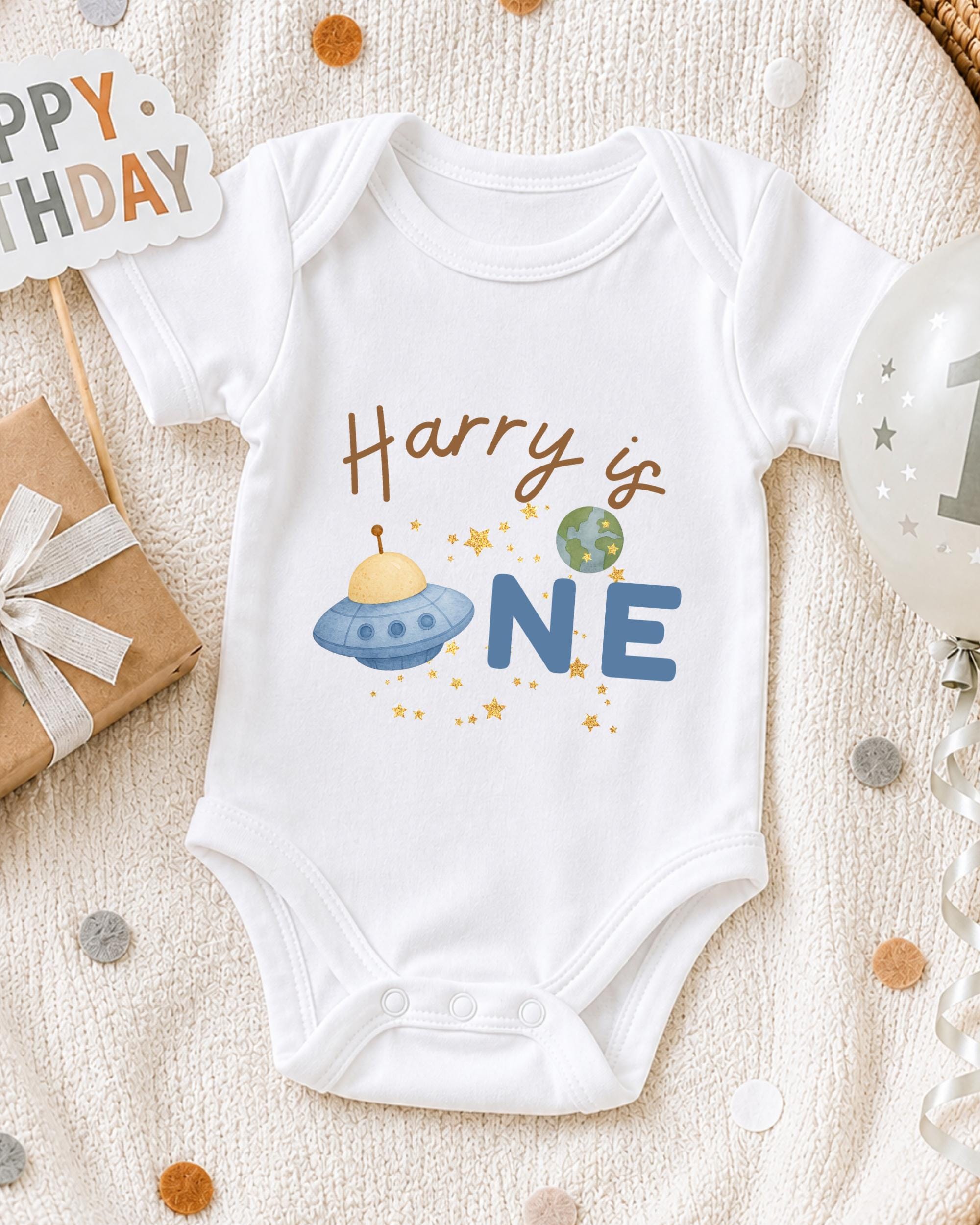 A white onesie with the text "Harry is one" printed on it, along with an illustration of a spaceship and a planet. The onesie is laid out on a blanket, surrounded by various party decorations such as a birthday sign, a balloon, and confetti.