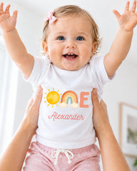 A smiling baby girl wearing a white shirt with a personalized name and a sun and rainbow design.