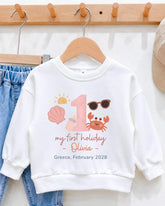 A white sweatshirt with a personalized design for a child's first holiday, featuring a sun, sunglasses, a shell, and a crab.