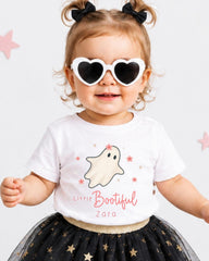 A young girl wearing a white t-shirt with a ghost design, heart-shaped sunglasses, and a black and gold tutu skirt, smiling and posing for the camera.