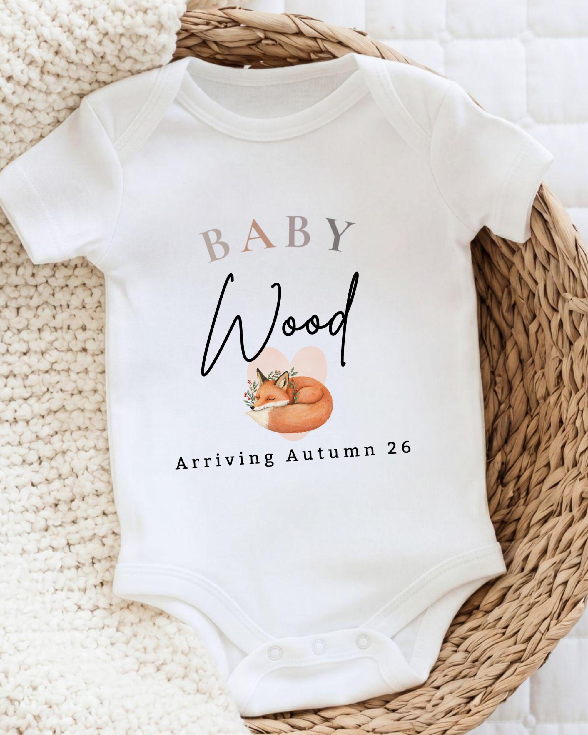A white baby onesie with the text "BABY Wood Arriving Autumn 26" printed on it, along with an illustration of a fox.
