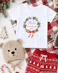 A white t-shirt with a Christmas wreath design, a beige knit hat with a bear face, red and white patterned shorts, and other holiday decorations are arranged on a plaid blanket.