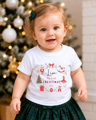 A smiling baby girl wearing a white t-shirt with a personalized design and text, standing in front of a Christmas tree.