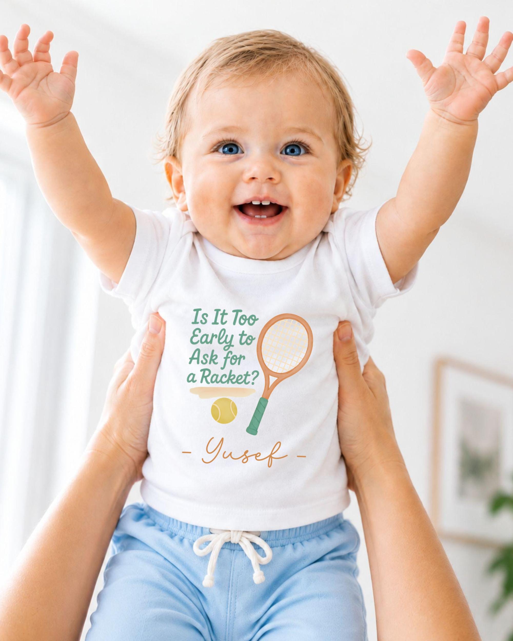 A smiling baby wearing a white shirt with a tennis racket and ball graphic, being held up by someone's arms.