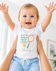 A smiling baby wearing a white shirt with a tennis racket and ball graphic, being held up by someone's arms.