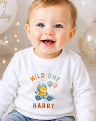 A smiling baby boy wearing a white shirt with a cartoon lion and the text "Wild One Harry" is the focus of the image.