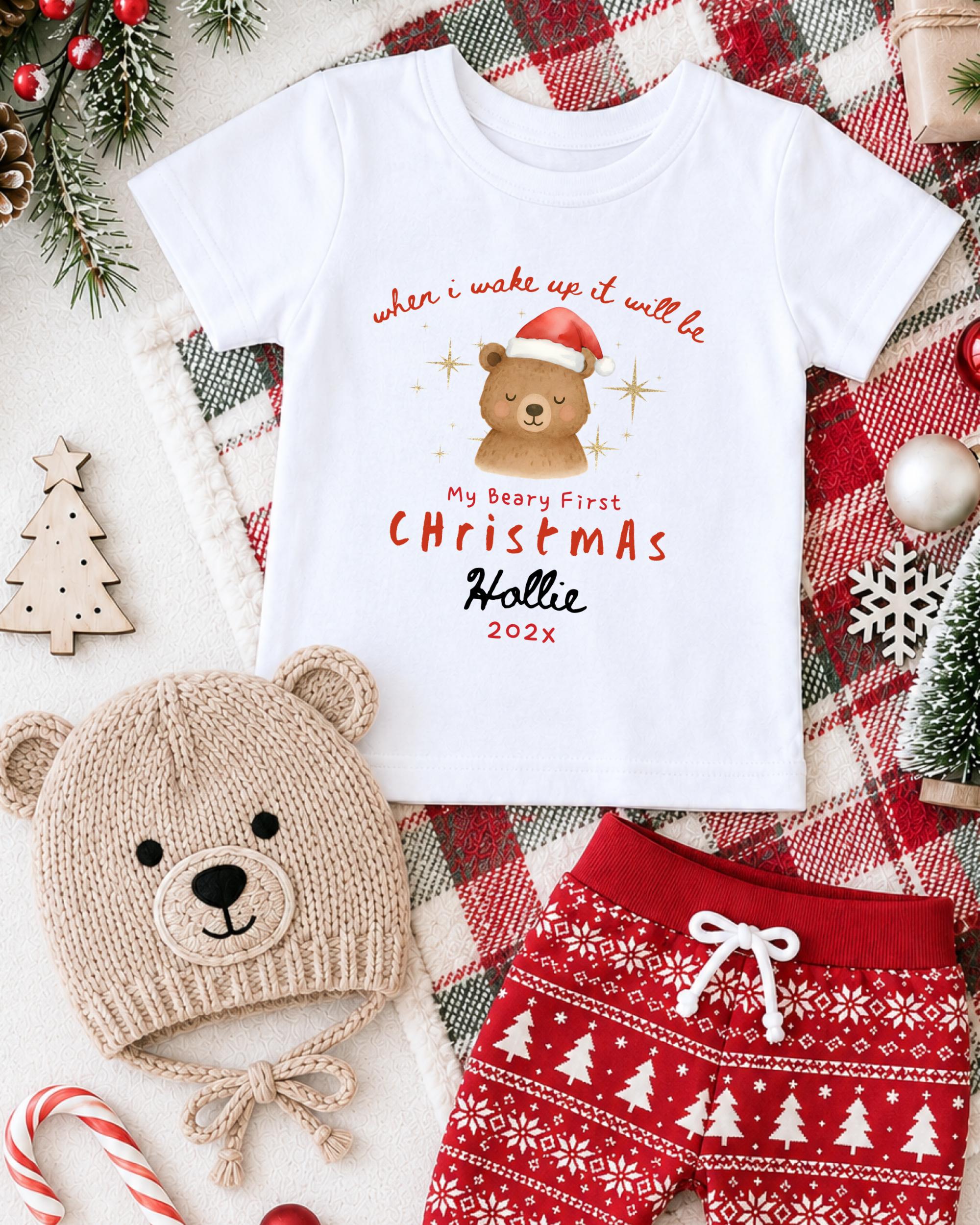 A white t-shirt with a Christmas-themed design, a knit hat with a bear's face, and red and white patterned shorts, all arranged on a plaid blanket with Christmas decorations.
