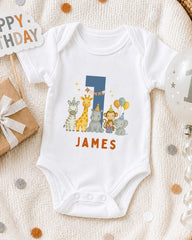 A white onesie with a cartoon image of a giraffe, monkey, and elephant, along with the name "James" printed on it, is displayed on a bed with a "Happy Birthday" sign and a balloon.