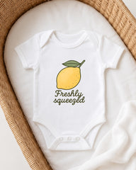 A white onesie with a lemon graphic and the text "Freshly squeezed" printed on it, placed on a white blanket inside a woven basket.