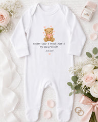 A white onesie with a teddy bear graphic and text, along with a pink ribbon, a ring, and some flowers, all arranged on a white background.