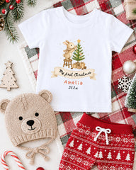 A white t-shirt with a Christmas-themed design, a beige knit hat, red shorts, and other Christmas-themed items are arranged on a plaid blanket.