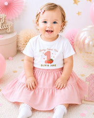 A smiling baby girl wearing a white t-shirt with a personalized design and a pink skirt, sitting on a pink and white background with balloons and confetti.