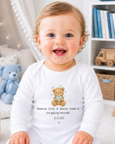 A smiling baby wearing a white onesie with a teddy bear graphic and text that reads "Auntie Lily & Uncle Josh's are getting married! 20.10.20".