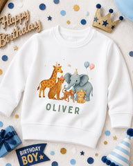 A white sweatshirt with a cartoon illustration of various animals, including a giraffe, elephant, and lion, along with the name "OLIVER" printed on it. The sweatshirt is surrounded by birthday decorations, including a crown, balloons, and confetti.