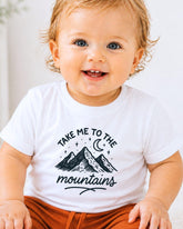 A smiling baby wearing a white t-shirt with a mountain graphic and the text "Take me to the mountains" printed on it.