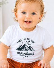 A smiling baby wearing a white t-shirt with a mountain graphic and the text "Take me to the mountains" printed on it.