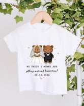 A white t-shirt with a graphic design of two teddy bears dressed as a bride and groom, along with the text "My Daddy & Mummy are getting married tomorrow!" and the date "20.10.2023".