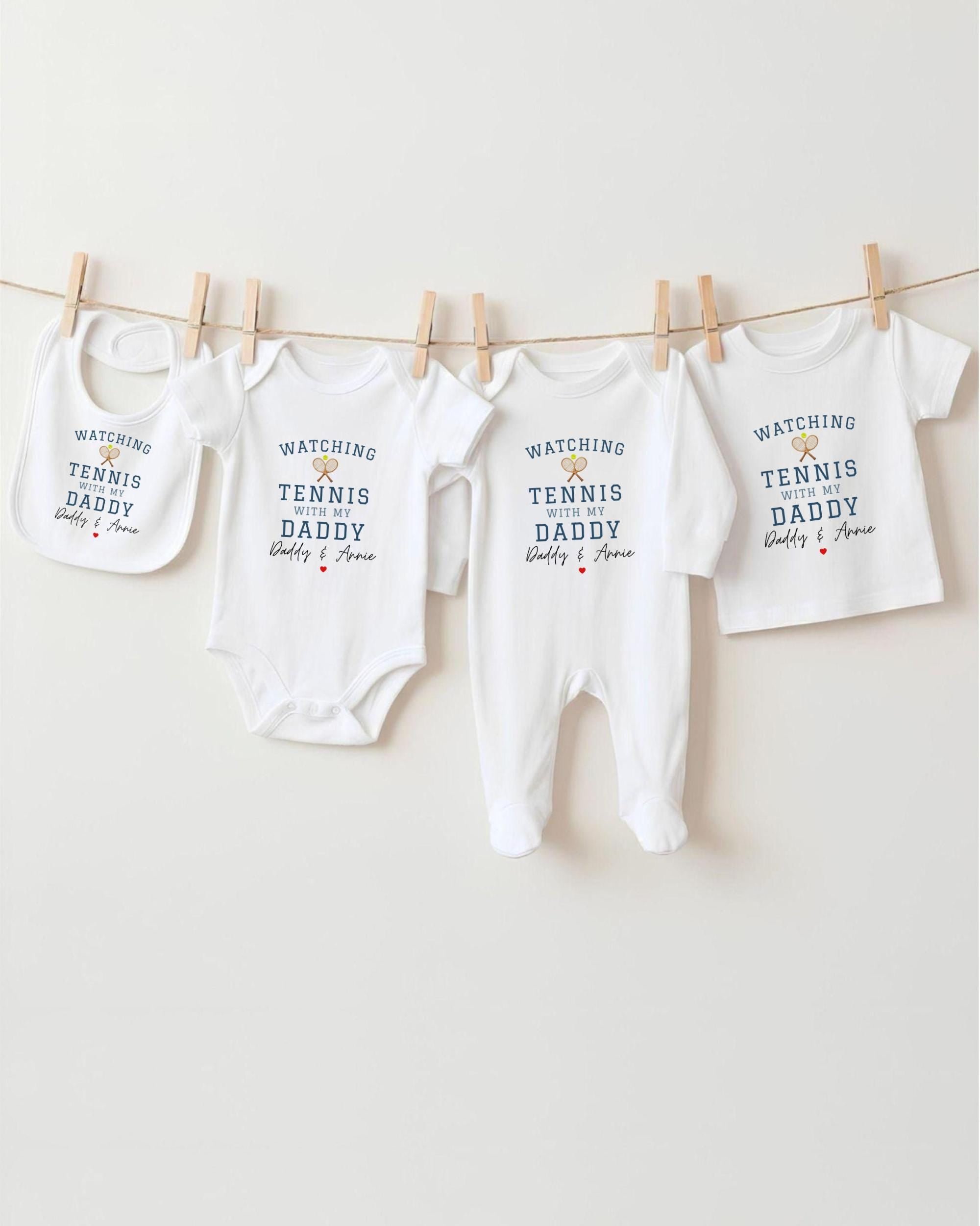 A row of four white baby clothes with the text "WATCHING TENNIS DADDY Daddy & Ava" printed on them, hanging on a clothesline.