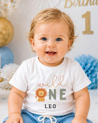 A smiling baby boy wearing a white t-shirt with a lion graphic and the text "wild one Leo" is sitting in front of a birthday party backdrop.