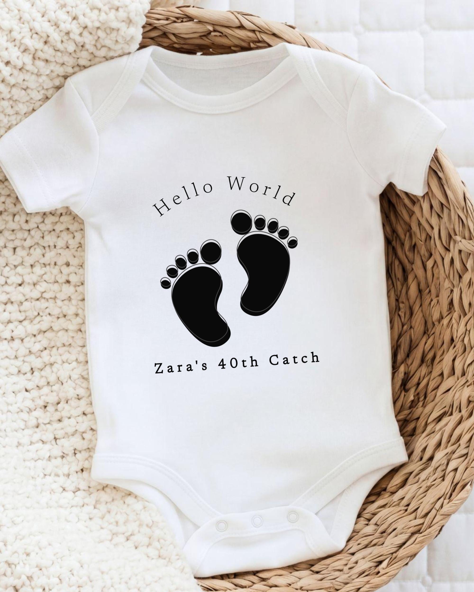 A white baby onesie with a black footprint design and text printed on it, placed on a woven basket.