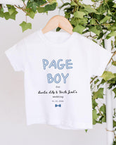 A white t-shirt with the text "PAGE BOY" printed on it, along with a date and a name. The t-shirt is hanging on a wooden hanger, and there are green leaves visible in the background.