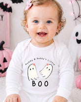 A smiling baby girl wearing a white onesie with a ghost design and text, sitting in front of Halloween decorations.