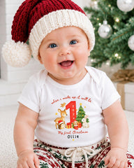 A smiling baby wearing a red and white Santa hat and a white t-shirt with a personalized design, sitting in front of a Christmas tree.