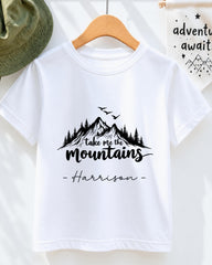 A white t-shirt with a mountain landscape design and the text "Take me the mountains Harrison" printed on it.
