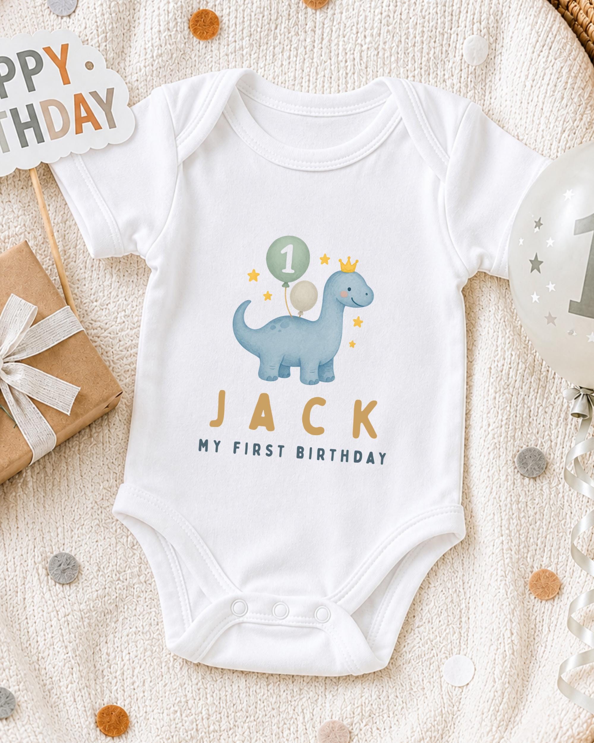 A white onesie with a dinosaur and the text "JACK MY FIRST BIRTHDAY" printed on it, along with a birthday sign and a balloon.