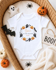 A white onesie with a Halloween-themed design, including bats, pumpkins, and a ghost, is displayed in a wicker basket. The onesie also has the text "Oscar, our first Halloween 2020" printed on it.