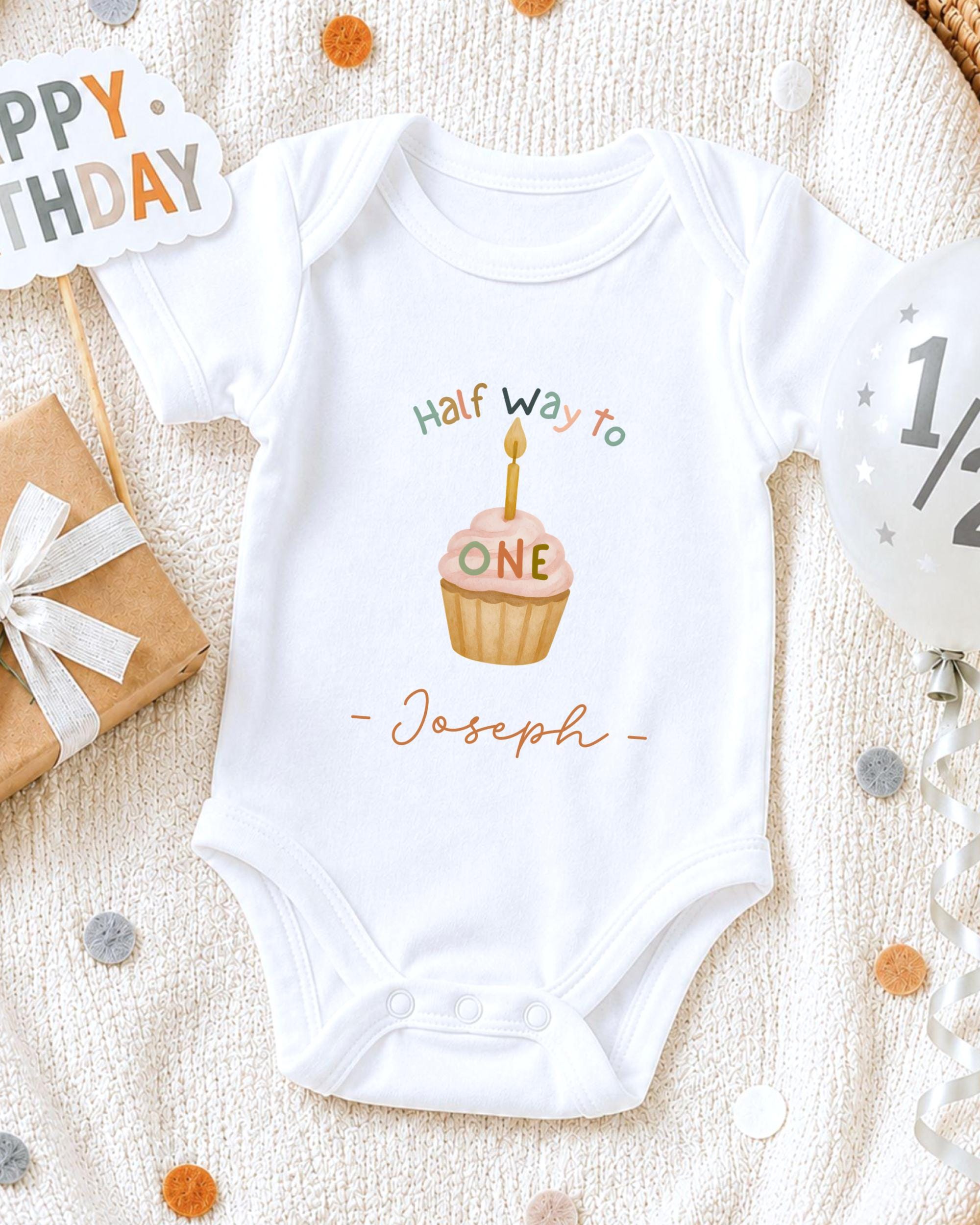A white onesie with a cupcake design and the text "Half way to Joseph" printed on it, along with a "Happy Birthday" sign and confetti.