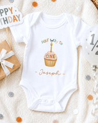 A white onesie with a cupcake design and the text "Half way to Joseph" printed on it, along with a "Happy Birthday" sign and confetti.