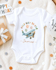 A white onesie with a cartoon airplane and the text "Half way to one - James" printed on it, along with a birthday sign and confetti.