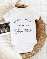 A white baby onesie with text announcing the arrival of a baby in October 2016, placed on a woven basket.