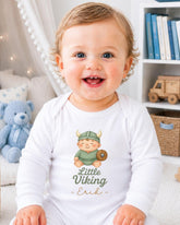 A smiling baby boy wearing a white onesie with a cartoon image of a Viking character and the text "Little Viking Eric" printed on it.