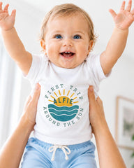 A smiling baby wearing a white shirt with a graphic design and text, being held up by someone's arms.