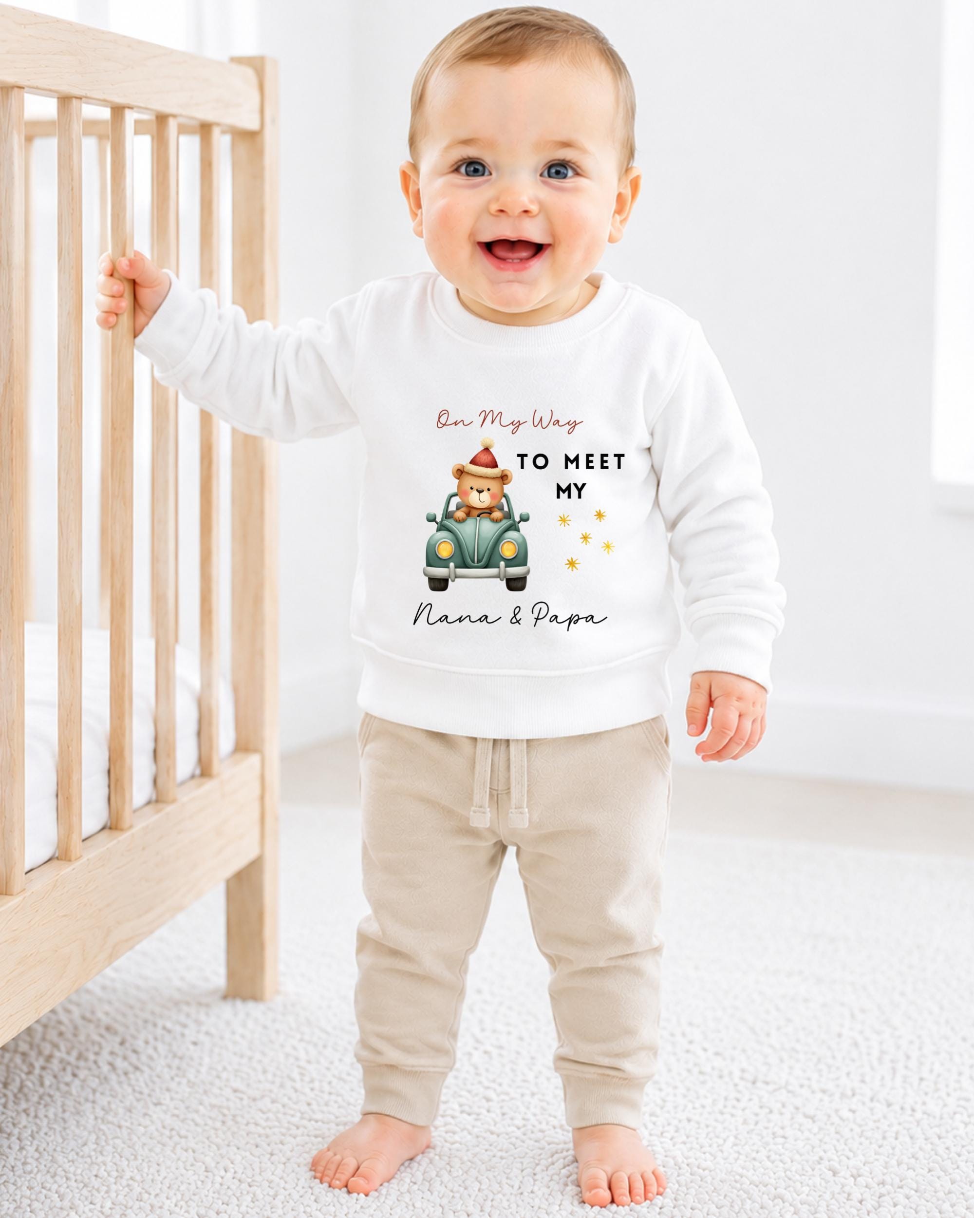 A smiling baby boy wearing a white sweatshirt with a cartoon image of a bear and the text "On My Way to Meet My Nana & Papa" standing in front of a wooden crib.