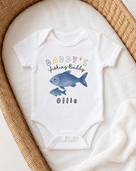 A white onesie with a fish design and the text "DADDY'S FISHING BUDDY OLLIE" printed on it, placed on a white blanket.
