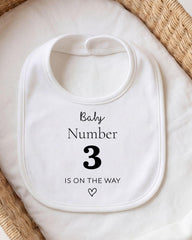 A white bib with the text "Baby Number 3 is on the way" printed on it.