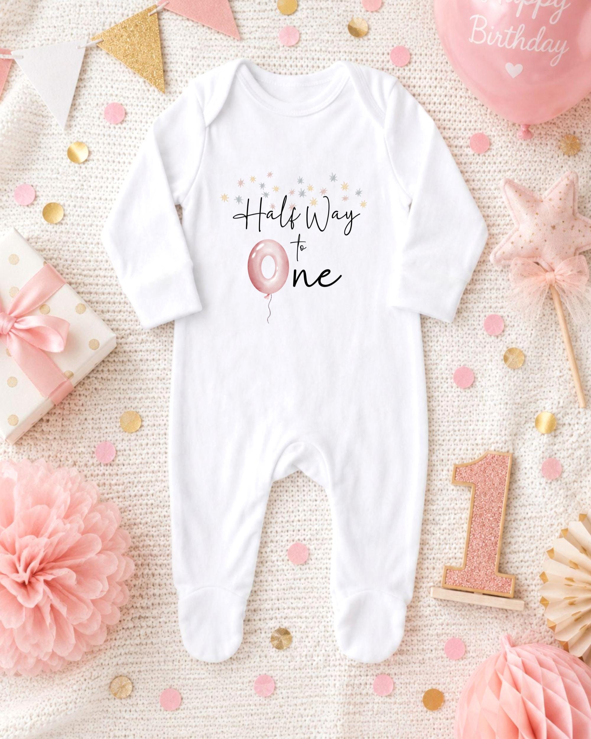 A white onesie with the text "Half Way to One" printed on it, surrounded by pink and gold confetti, a pink balloon, and other baby-themed decorations.