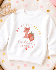A white sweatshirt with a graphic design featuring a strawberry and the text "My Berry First birthday Jessica" printed on it.